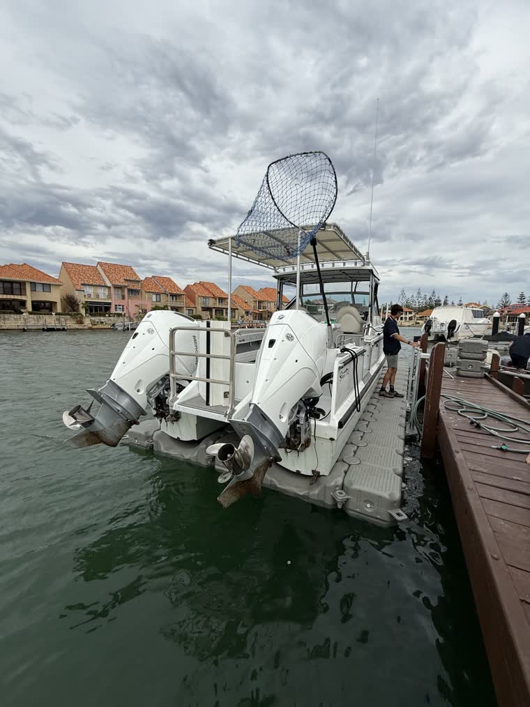 A boat with two engines and a net on a pole on the back docked on a cloudy day with a person standing next to the boat